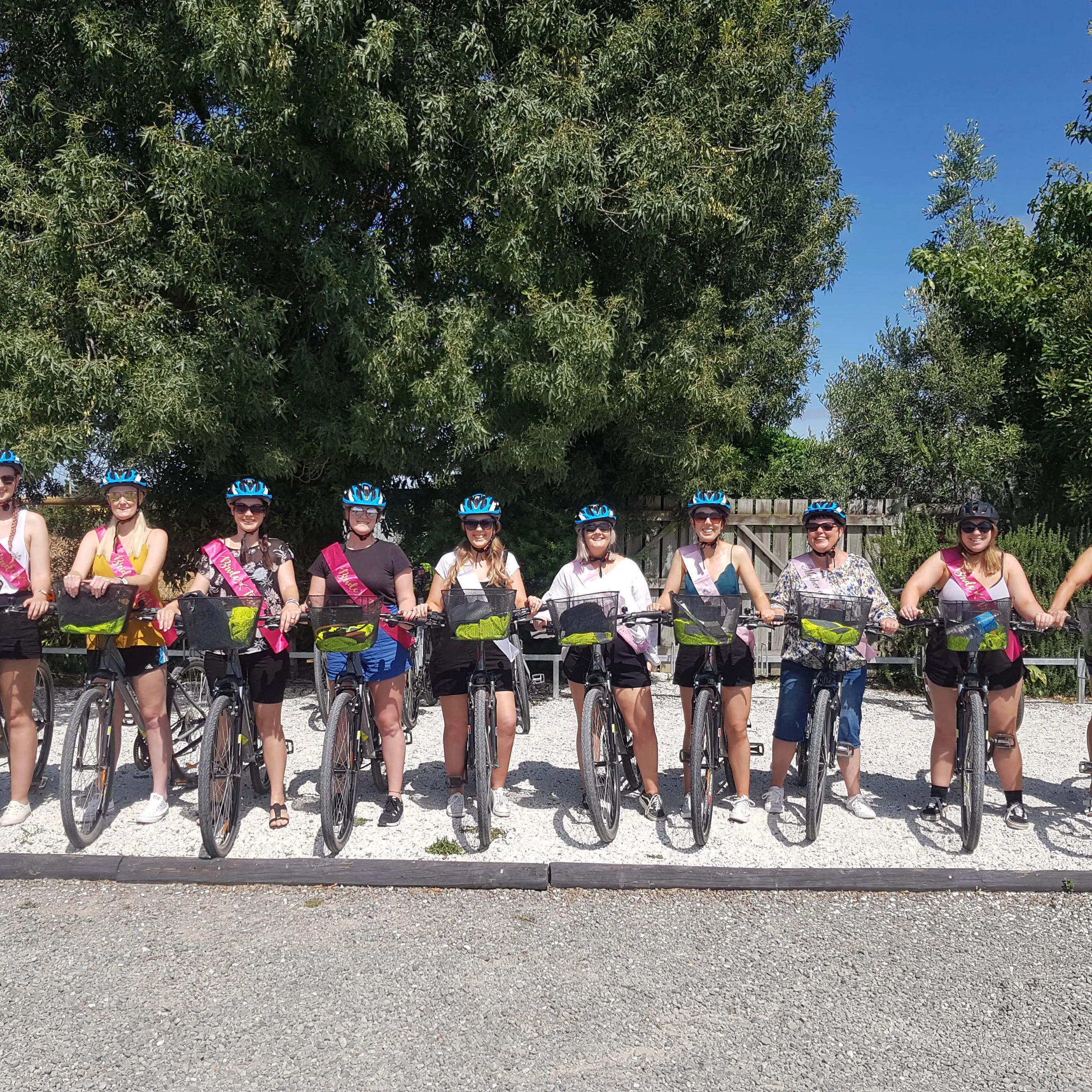 Group of cyclists posing with bikes on gravel path, wearing helmets and sashes, trees in background.