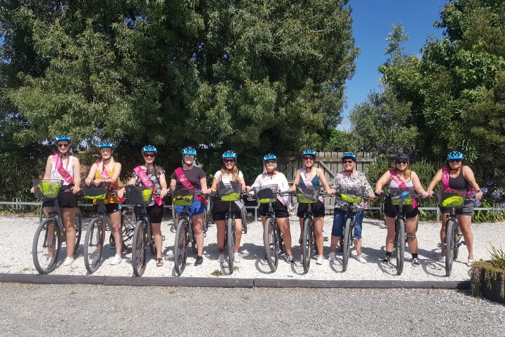 Group of cyclists posing with bikes on gravel path, wearing helmets and sashes, trees in background.