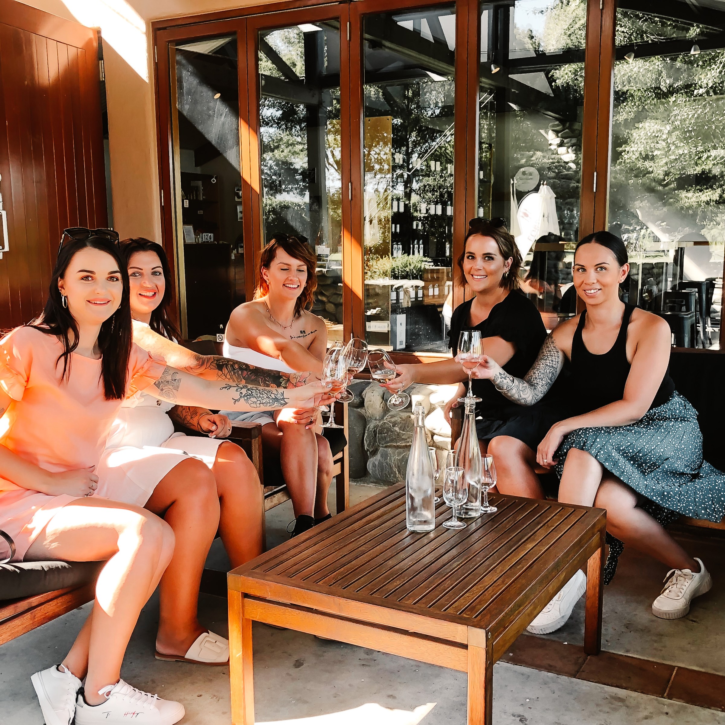 Five women sitting on patio furniture toasting with wine glasses outside a Marlborough Winery.