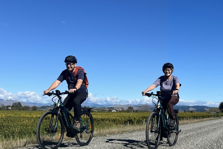 Two cyclists on a gravel road with fields and mountains under a clear Marlborough blue sky.