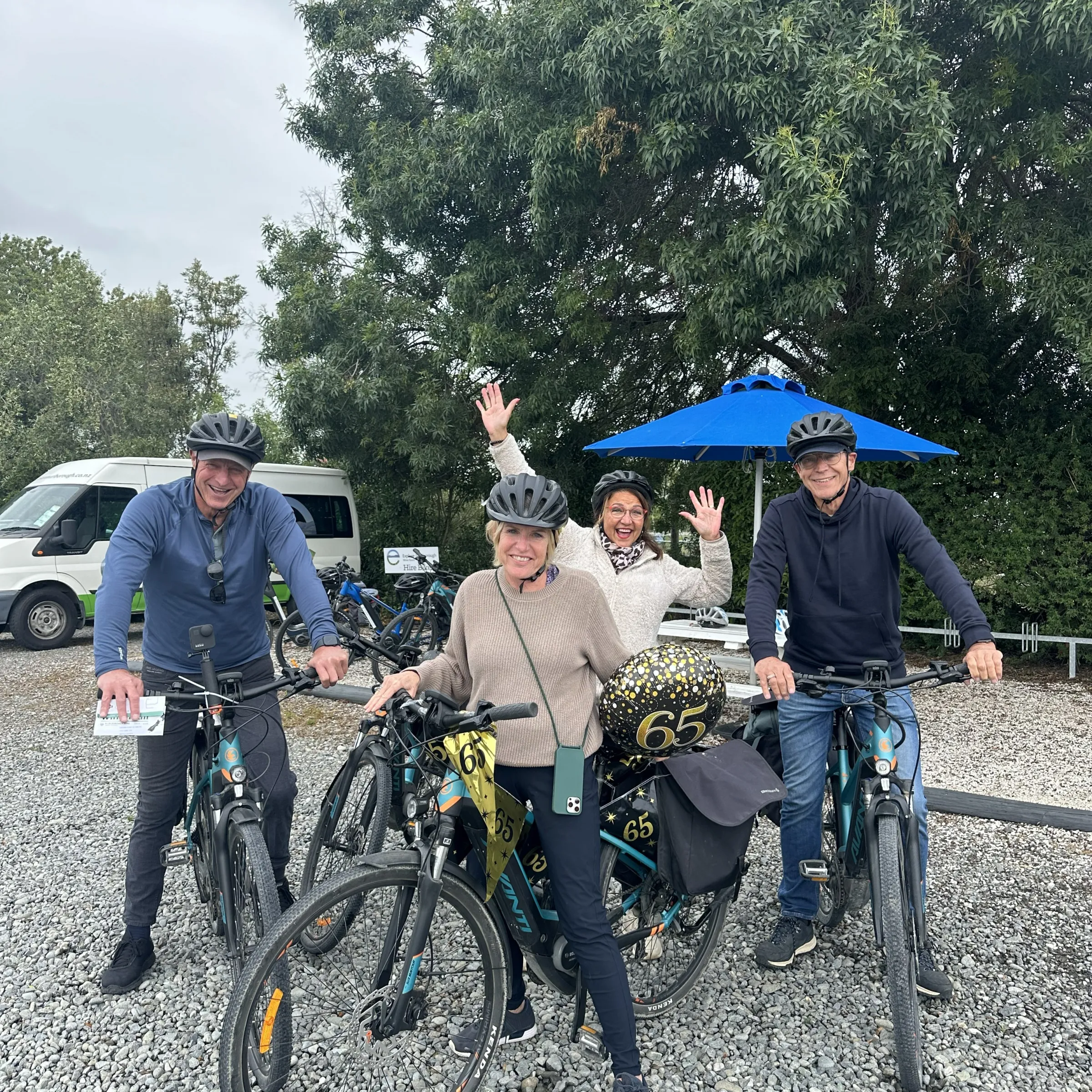 Four people on bikes celebrating with '65' balloons and helmets, standing on gravel.