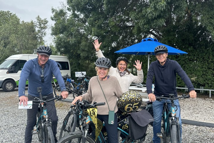 Four people on bikes celebrating with '65' balloons and helmets, standing on gravel.
