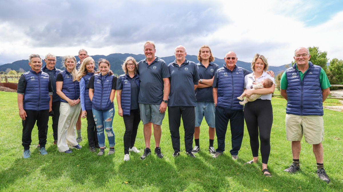 A group of twelve people and a baby standing on grass with mountains in the background.
