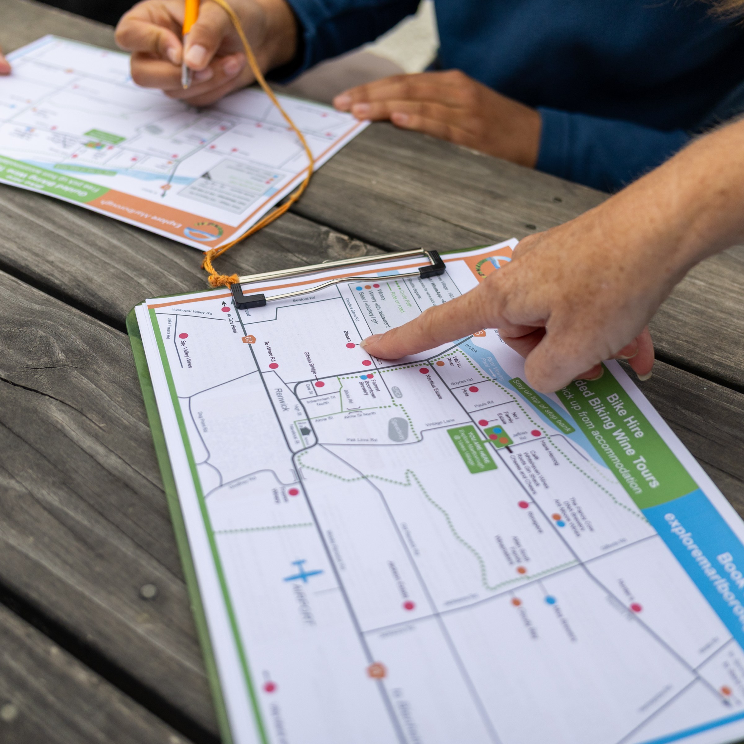 Two people examine a map on a wooden table, one pointing and one writing.