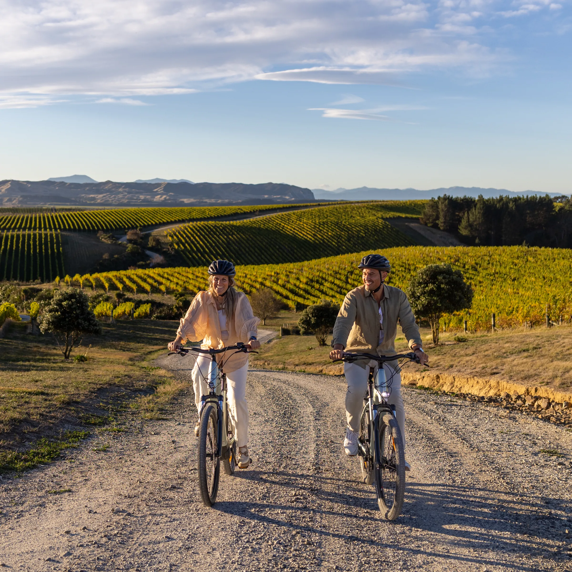 Two people cycling on a path through a vineyard with hills in the background.